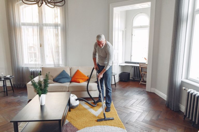 A man using a vacuum cleaner in a well-organized living room, with a sofa and coffee table in the background.